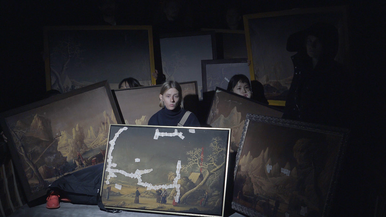 Still from video featuring several somber people in a darkened environment, each holding a large antique landscape painting, some paintings have tape over the canvas in places where they have been damaged