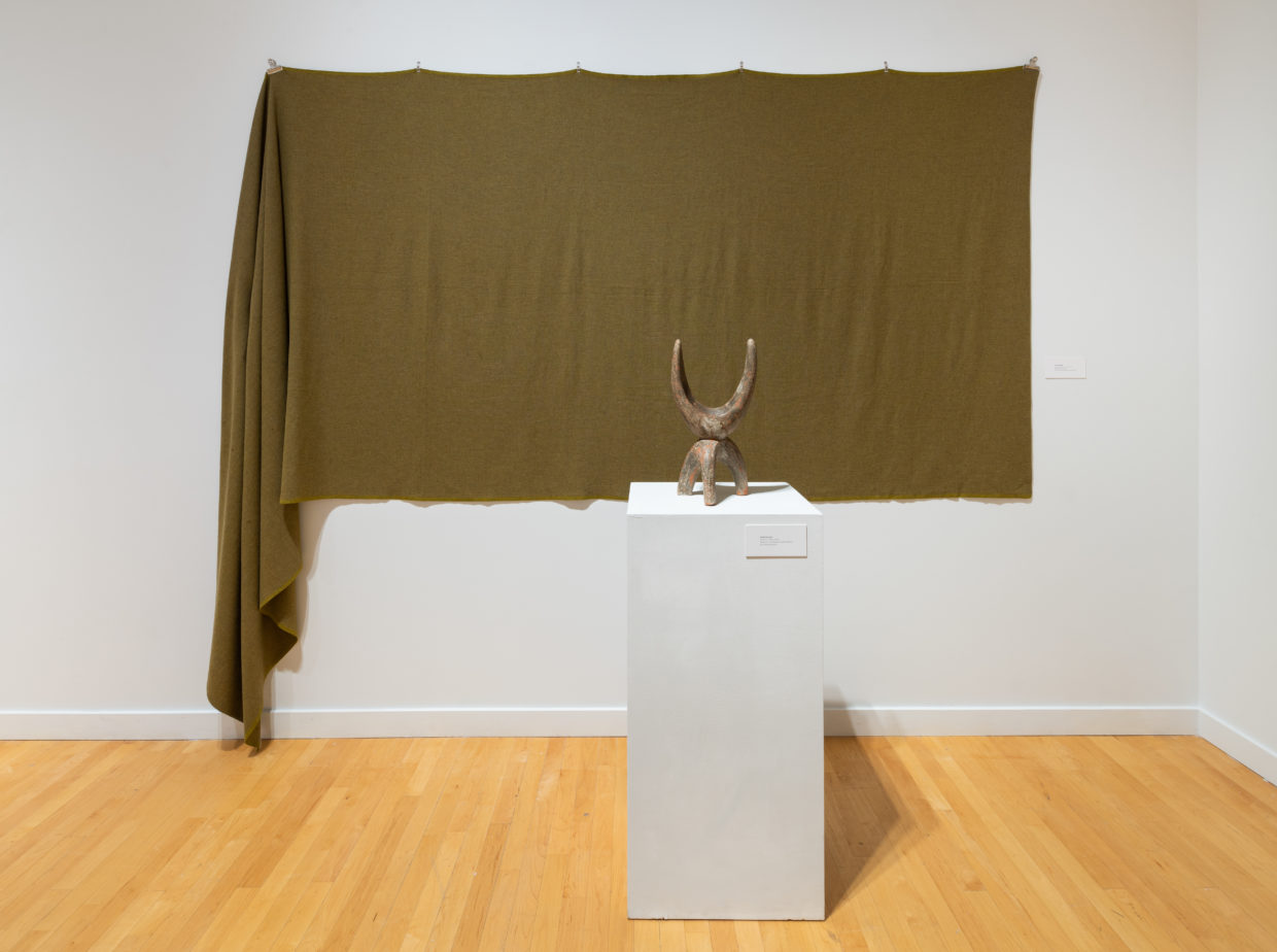 View of a gallery wall with a bolt of moss-green woven wool fabric hanging from clips, one side is draping down to the floor. In front, on a white pedestal, is a small beige ceramic sculpture that has a tripod-like base and a crescent-moon shaped upper that resembles animal horns.