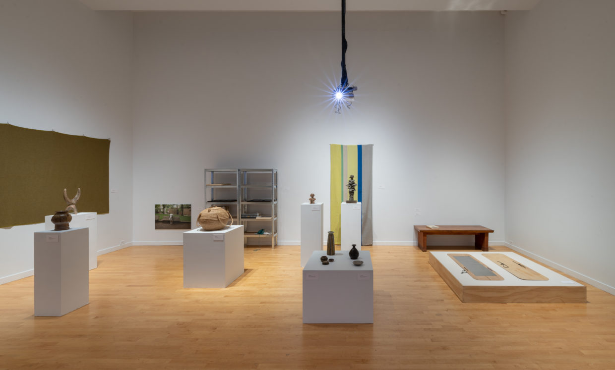 A white-walled gallery with several pedestals of varying heights displaying ceramics and a basket, woven wall hangings, flat artworks on a low dais, utility shelves, and a ceiling-mounted projector