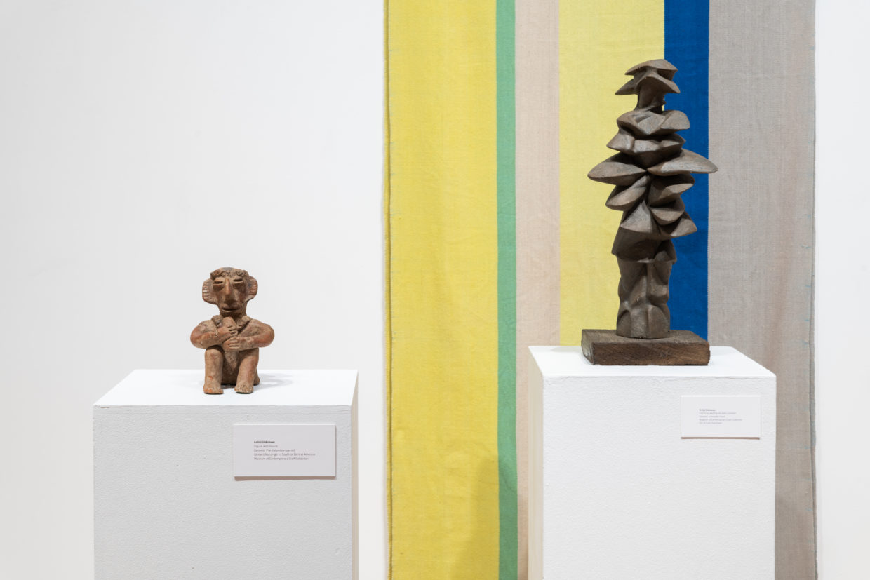 Two ceramic sculptures on pedestals in front of a woven tapestry in wide stripes of muted yellow, blue, gray, and green, One sculpture is a terracotta seated figurine with large ears that appears to be playing a flute, the other is an abstract stack of flat, pebble-like forms that vaguely resembles a human figure