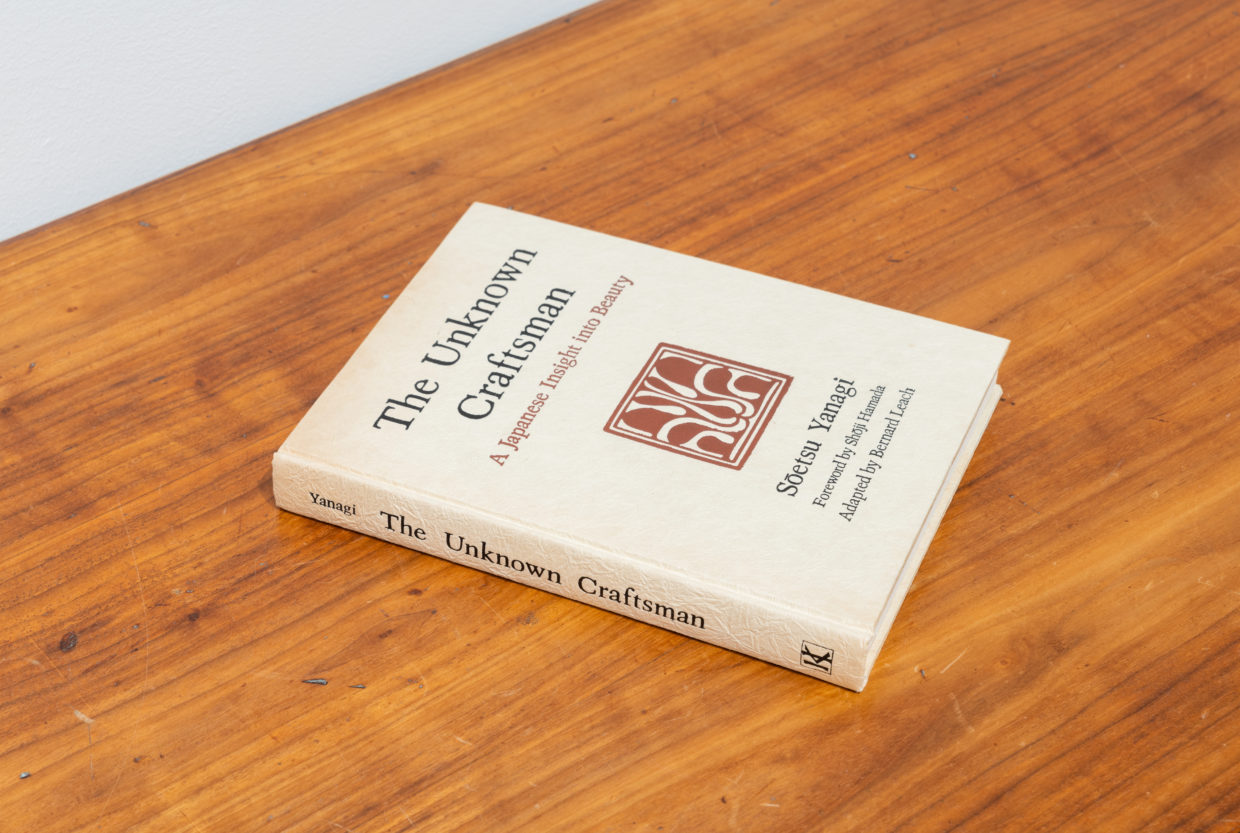 A vintage hardcover book with black lettering and a red insignia on off-white paper on a wooden bench