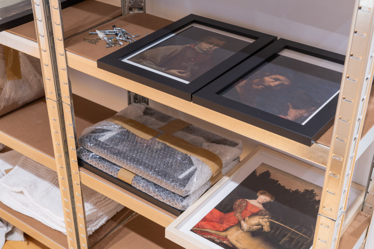 A wood and metal utility shelf with framed pictures of antique European paintings lying flat on shelves, some stacked and packaged in bubble wrap. A small pile of screws and other hanging fixtures is also on the shelf.