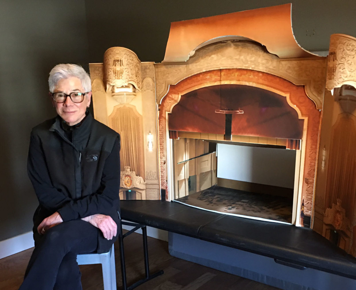 Artist Rose Bond seated in front of a scale model of the Arlene Schnitzer Concert Hall, Bond wears black athletic gear, black-rimmed glasses, and close cropped light gray hair, the model concert hall sits on a folding table in the gray room